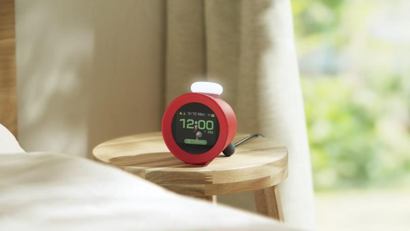 A Nintendo Alarmo clock is pictured on a bedside table in a sunlit room, displaying the time (12:00pm) and an animation of Olimar and a red Pikmin from the Pikmin franchise
